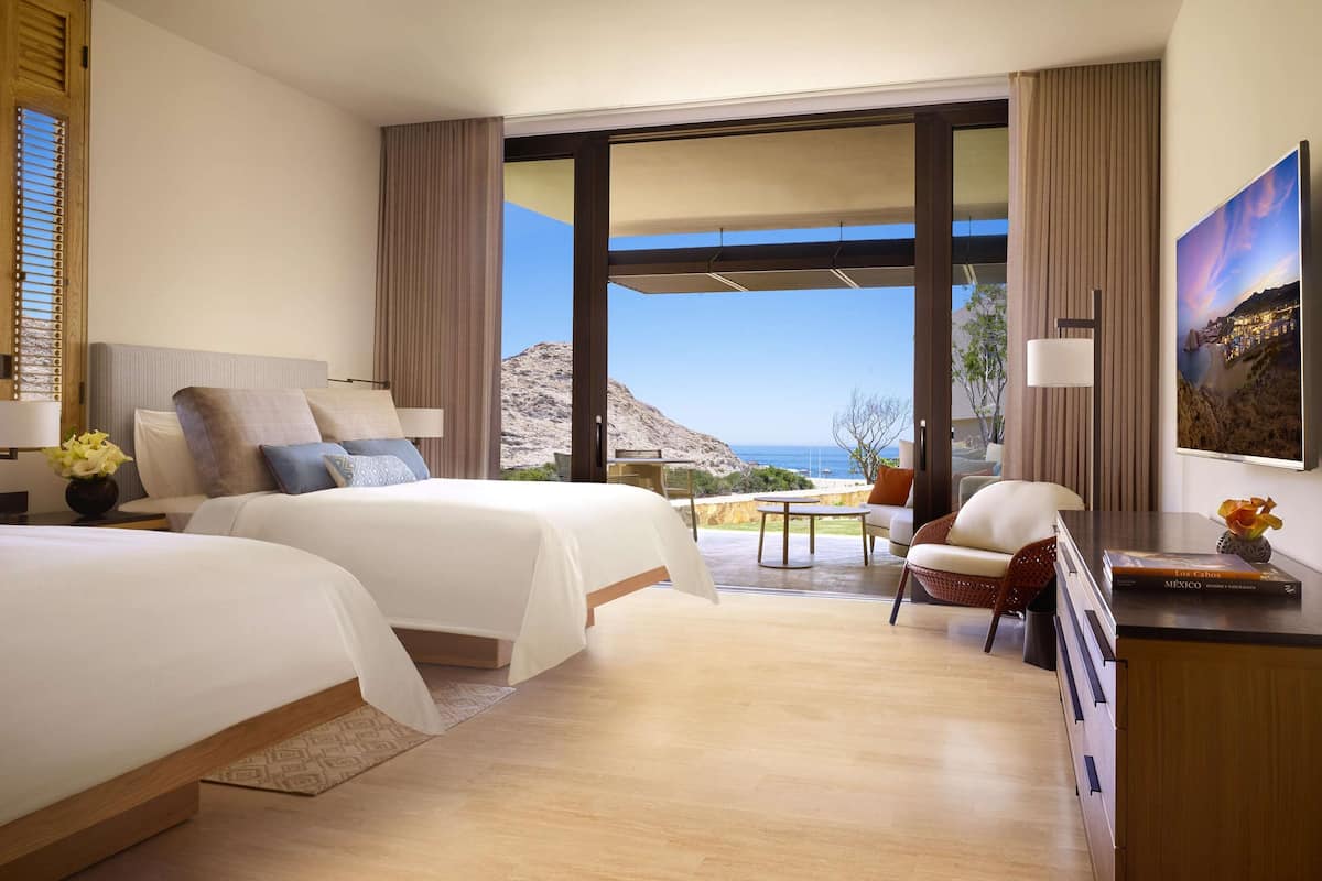 A hotel room with two beds, a desk, a chair, and a large window offering a view of the sea.