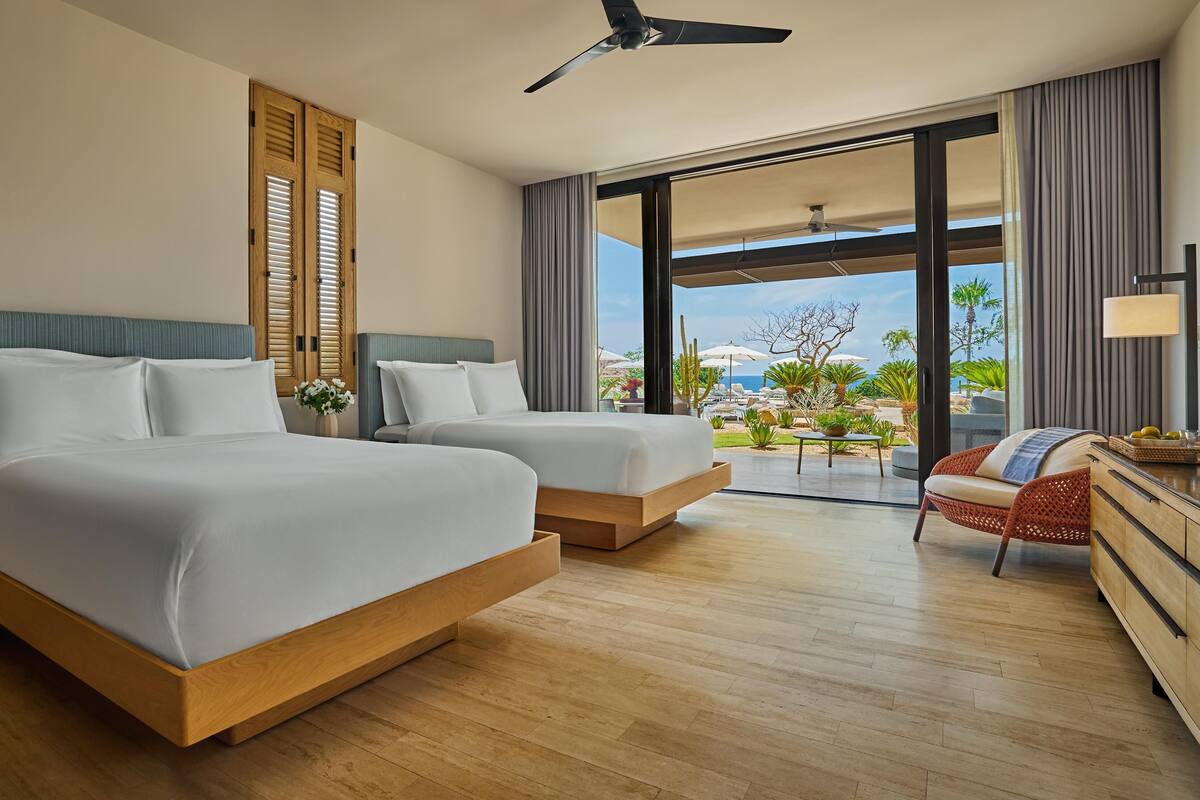 A hotel room with two beds, a ceiling fan, a view of the ocean, and a balcony with outdoor furniture.
