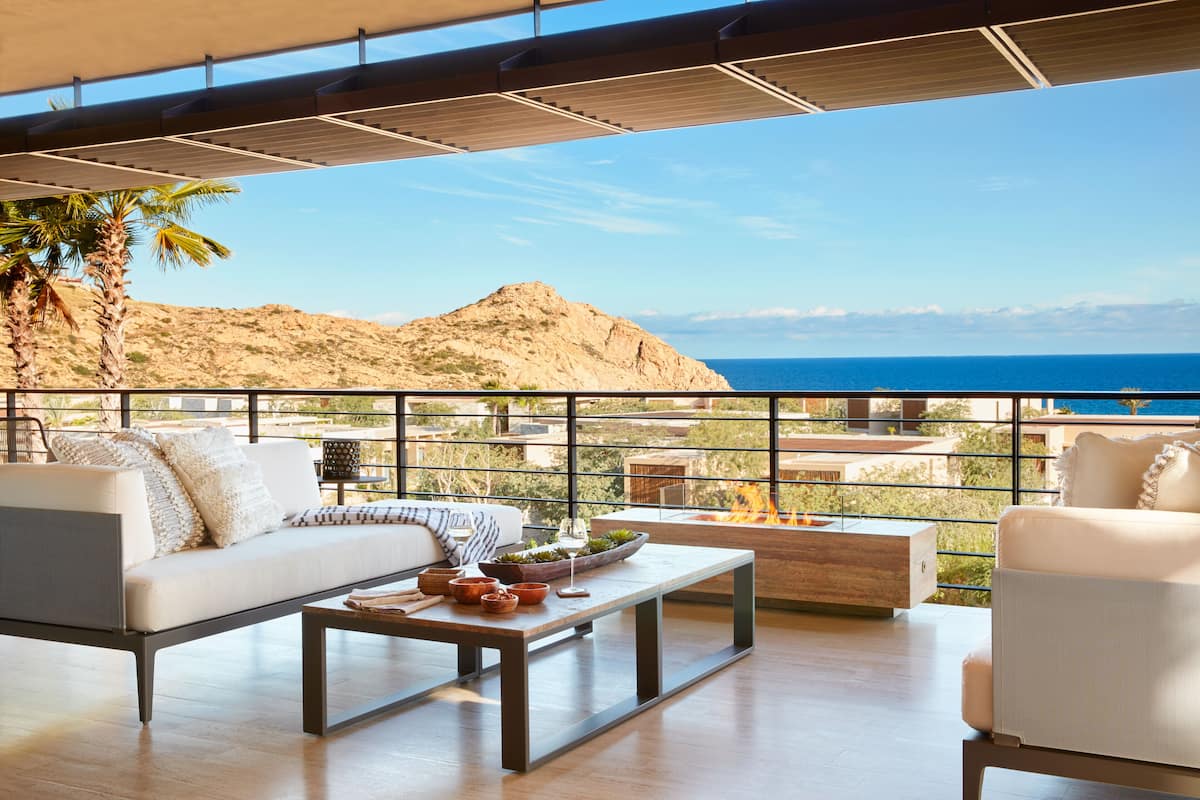 A modern outdoor living area with a sofa, armchairs, and a coffee table, overlooking a mountainous landscape and the sea.