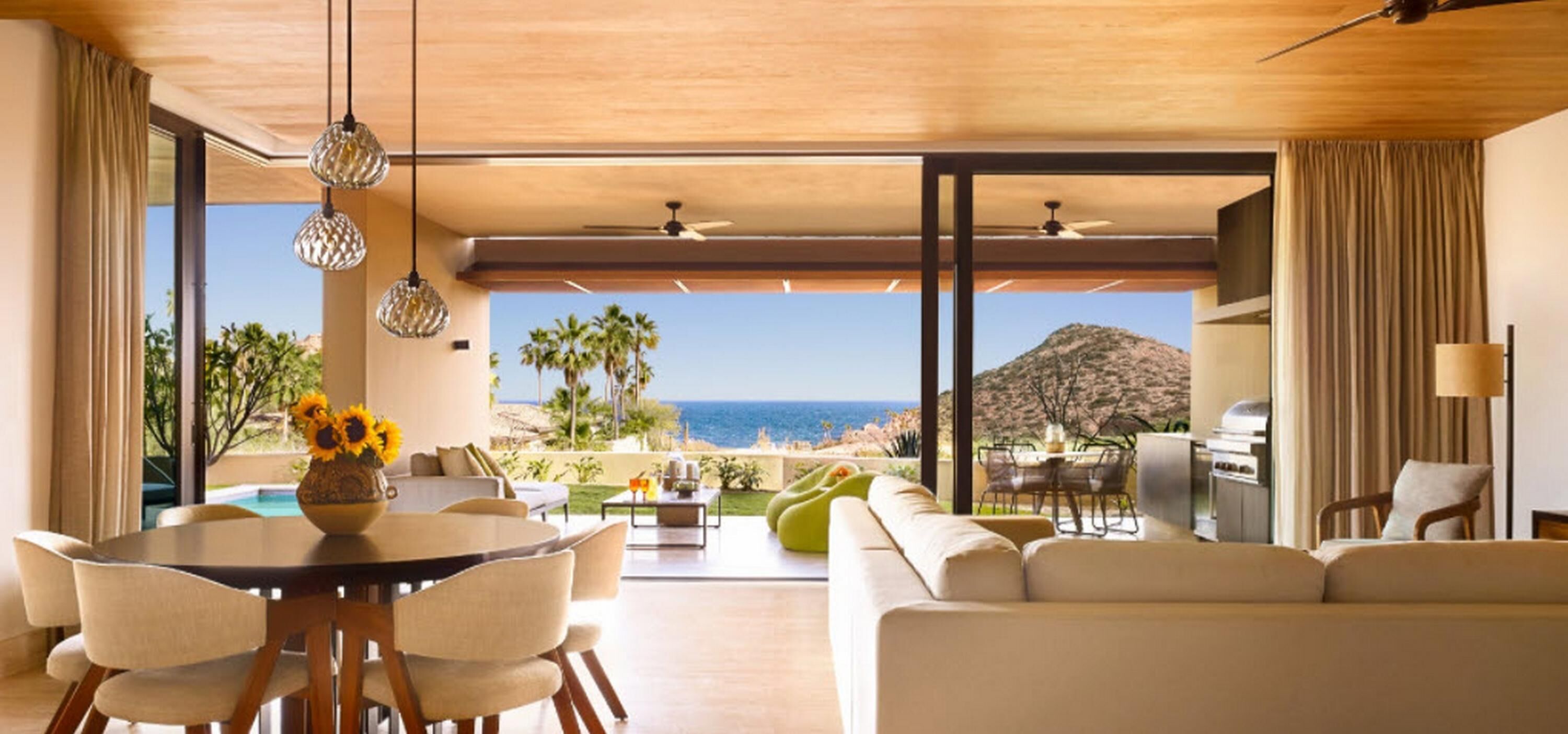 A modern living room with a large window overlooking a beach and ocean, furnished with a sofa, dining table, and chairs.