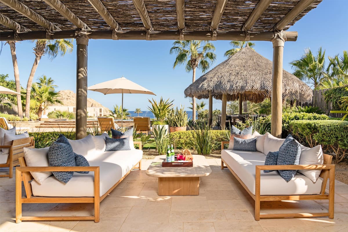 A beachside lounge area with wooden furniture, thatched umbrellas, and palm trees.