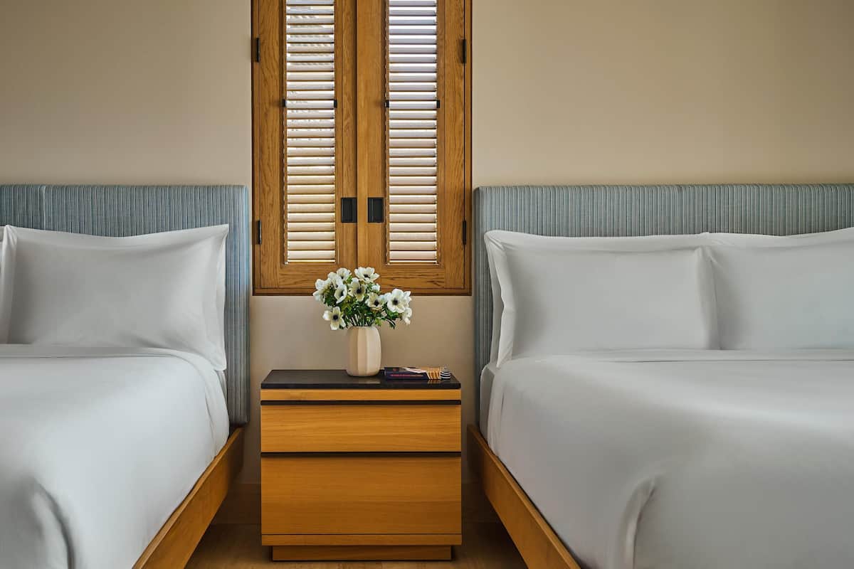 A hotel room with two beds, a wooden nightstand, and a window with wooden shutters.