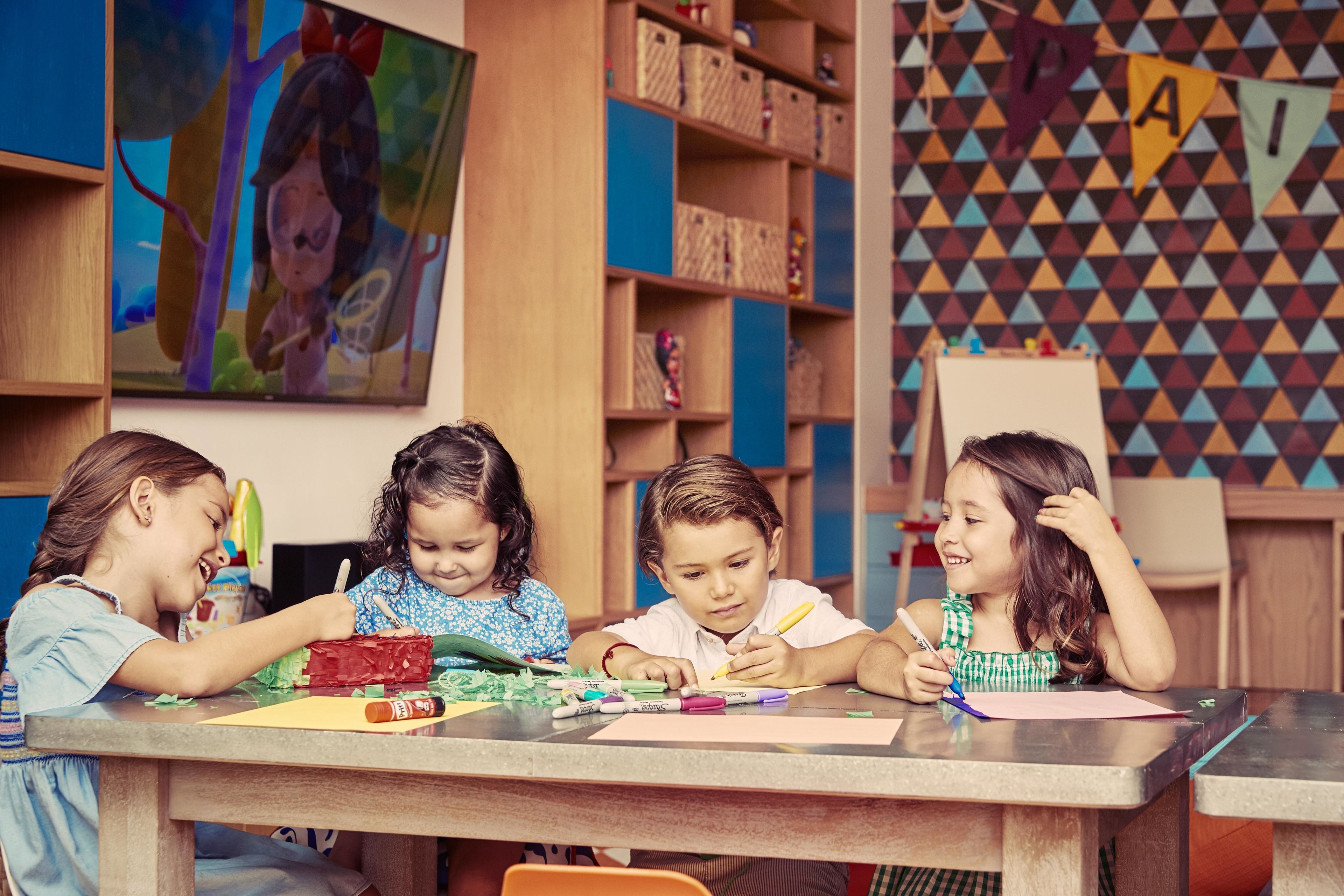 Un aula con niños realizando actividades artísticas, estanterías de madera con cajas de almacenamiento y un patrón de pared geométrico y colorido.