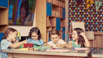 Un aula con niños realizando actividades artísticas, estanterías de madera con cajas de almacenamiento y un patrón de pared geométrico y colorido.