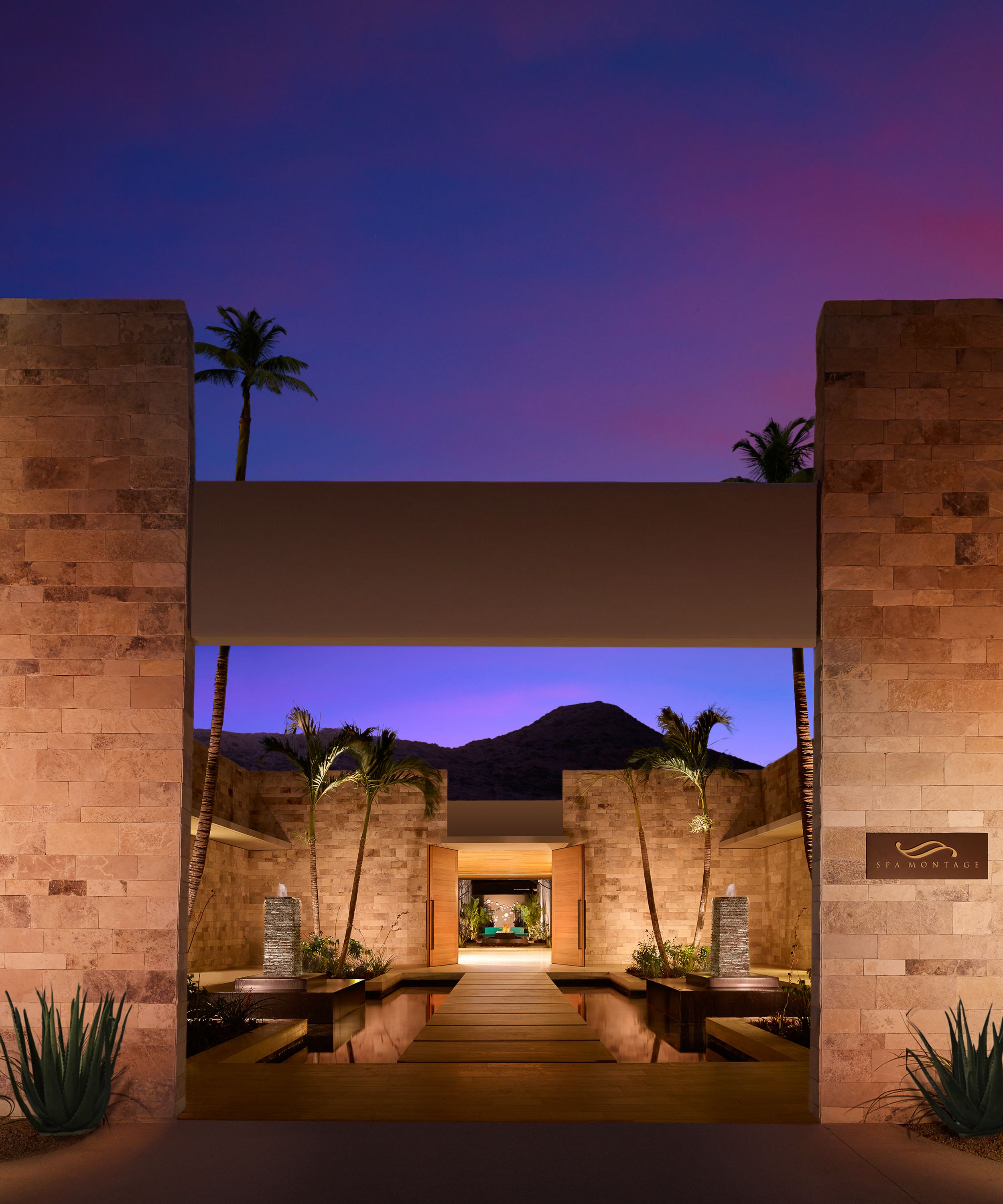 A modern spa entrance with a wooden pathway leading to a reception area, flanked by palm trees and stone pillars.