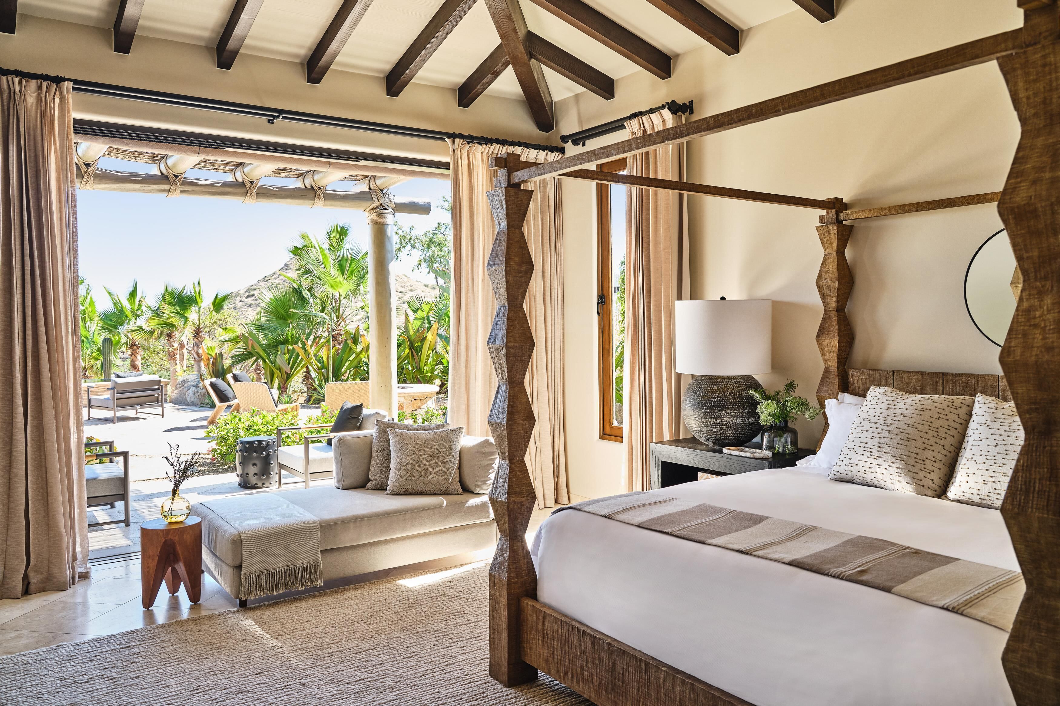 A spacious bedroom with a four-poster bed, a sitting area with a sofa and chairs, and a view of a tropical garden.