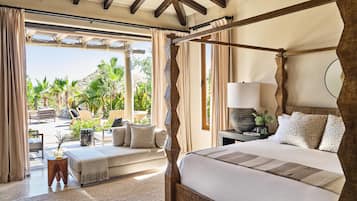 A spacious bedroom with a four-poster bed, a sitting area with a sofa and chairs, and a view of a tropical garden.