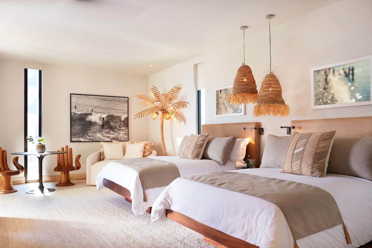A modern hotel room with two beds, a large window, a dining area, and a tropical plant.
