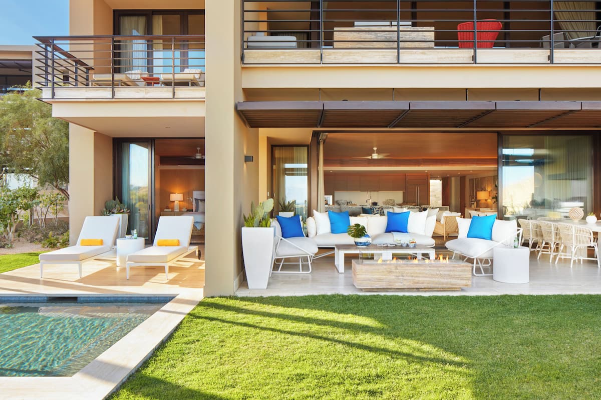A modern two-story residential building with a spacious outdoor living area featuring a pool, lounge chairs, and a dining area with white furniture and blue cushions.