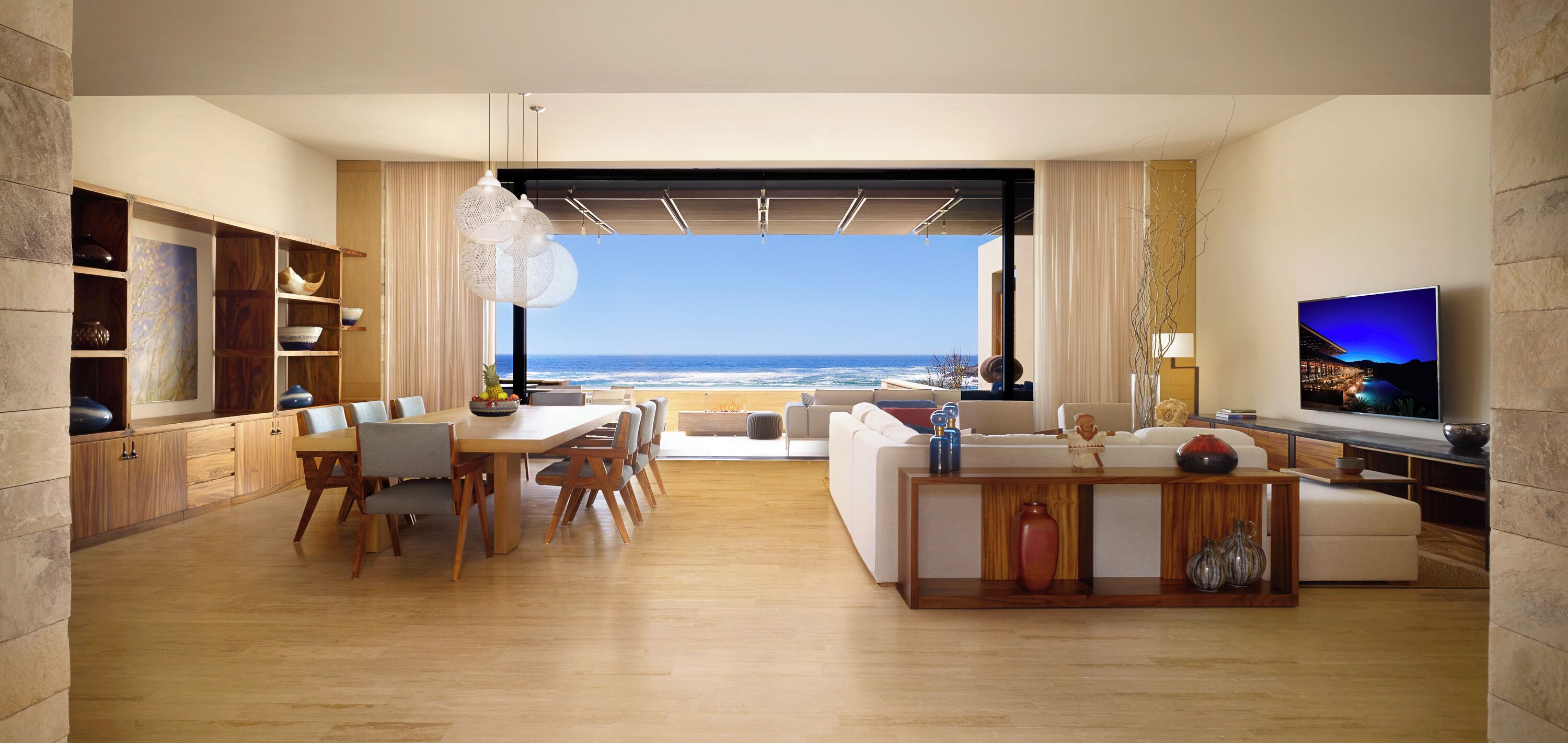 Una sala de estar moderna con un ventanal grande con vistas al océano, un comedor con muebles de madera y un televisor de pantalla plana sobre un pedestal de madera.
