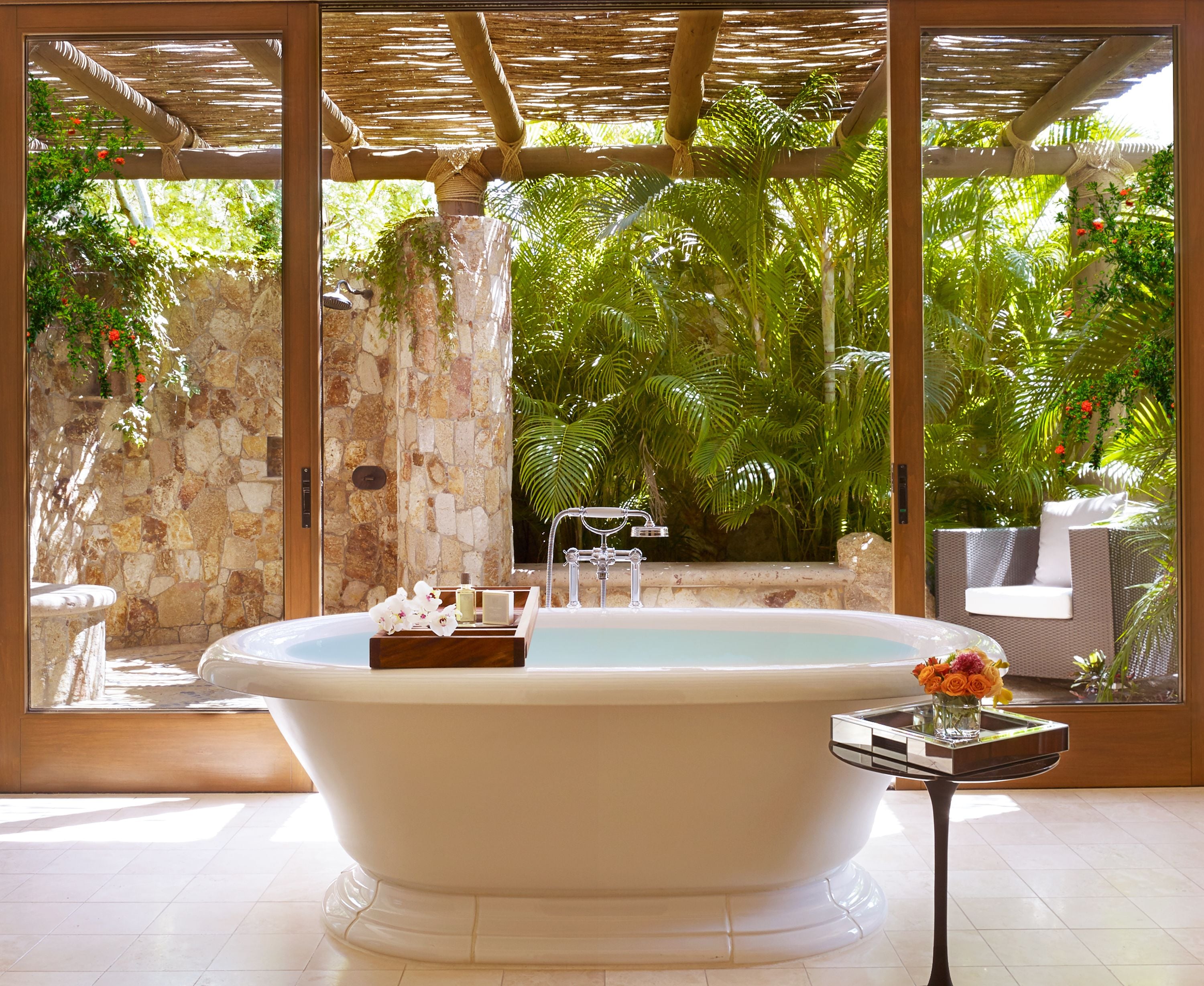 A large freestanding bathtub in a sunlit room with a stone wall and a view of lush greenery outside.