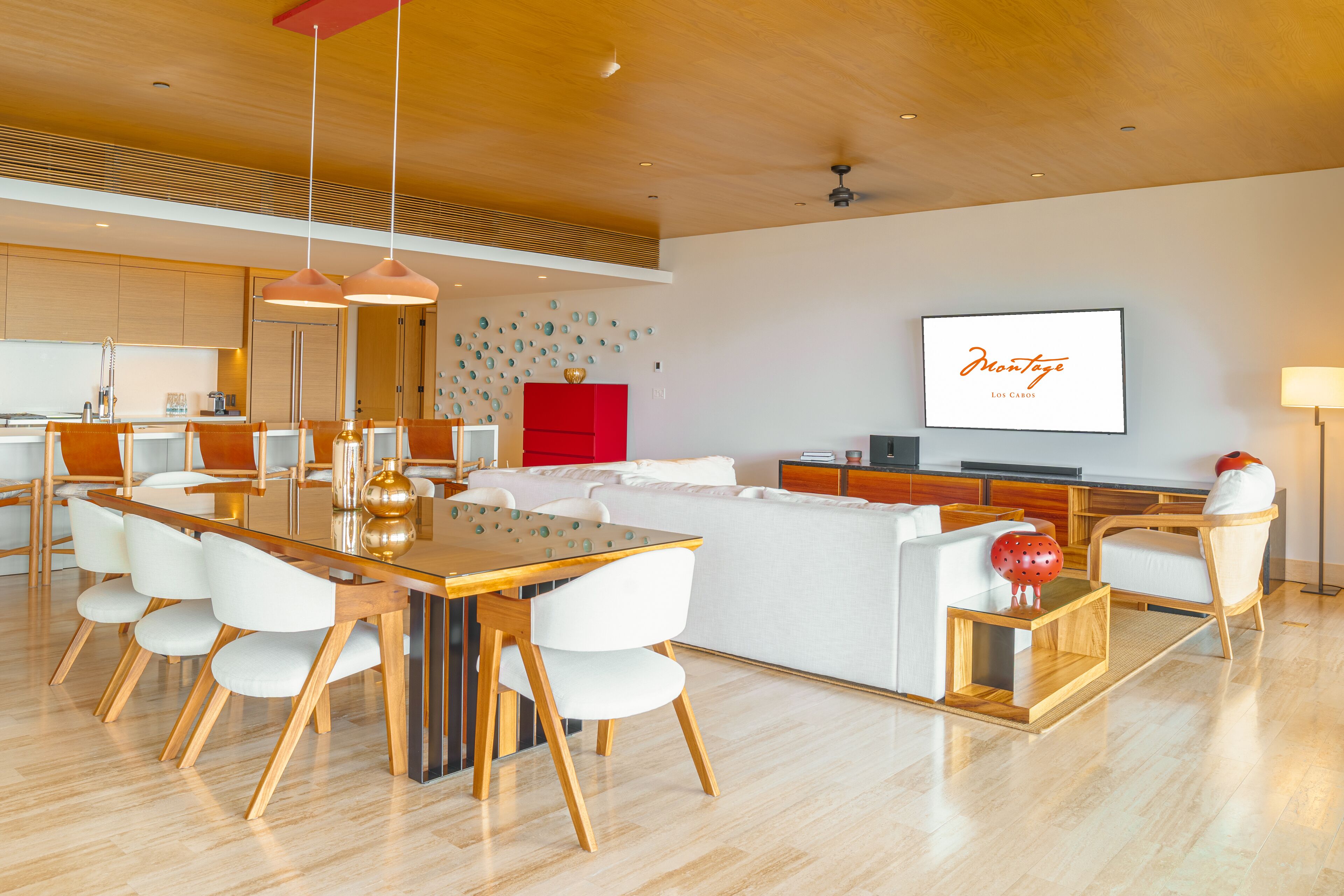 A modern living room with a wooden dining table, white chairs, a red accent wall, and a flat-screen TV.