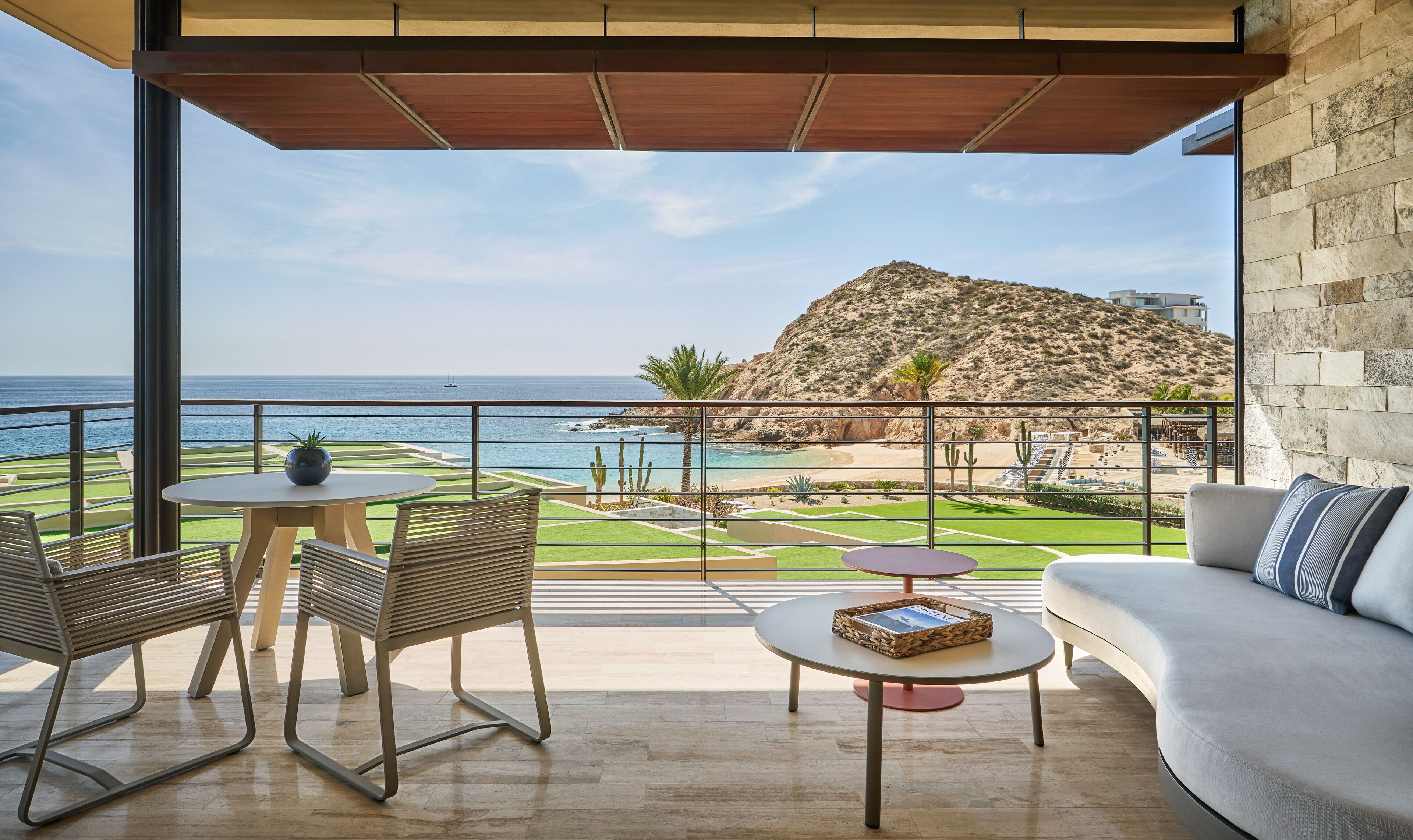 A balcony with a view of the sea and a mountain, furnished with a sofa, chairs, and a table.
