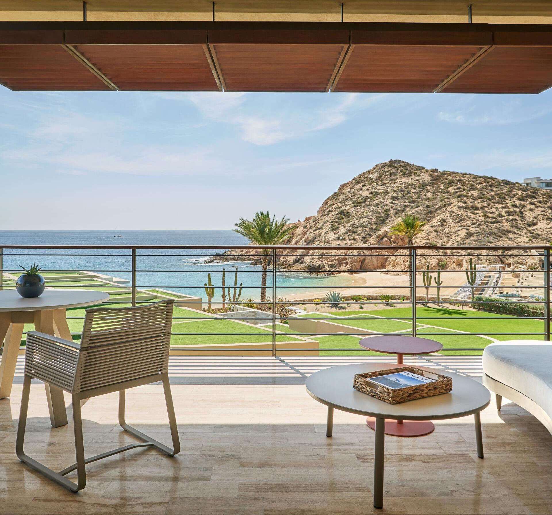 A balcony with a view of the sea and a hill. There are chairs and a table on the balcony.