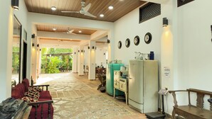 Una habitación amplia con techo de madera, ventilador de techo y suelo de baldosas. Hay varios refrigeradores de estilo antiguo y un banco con equipaje.