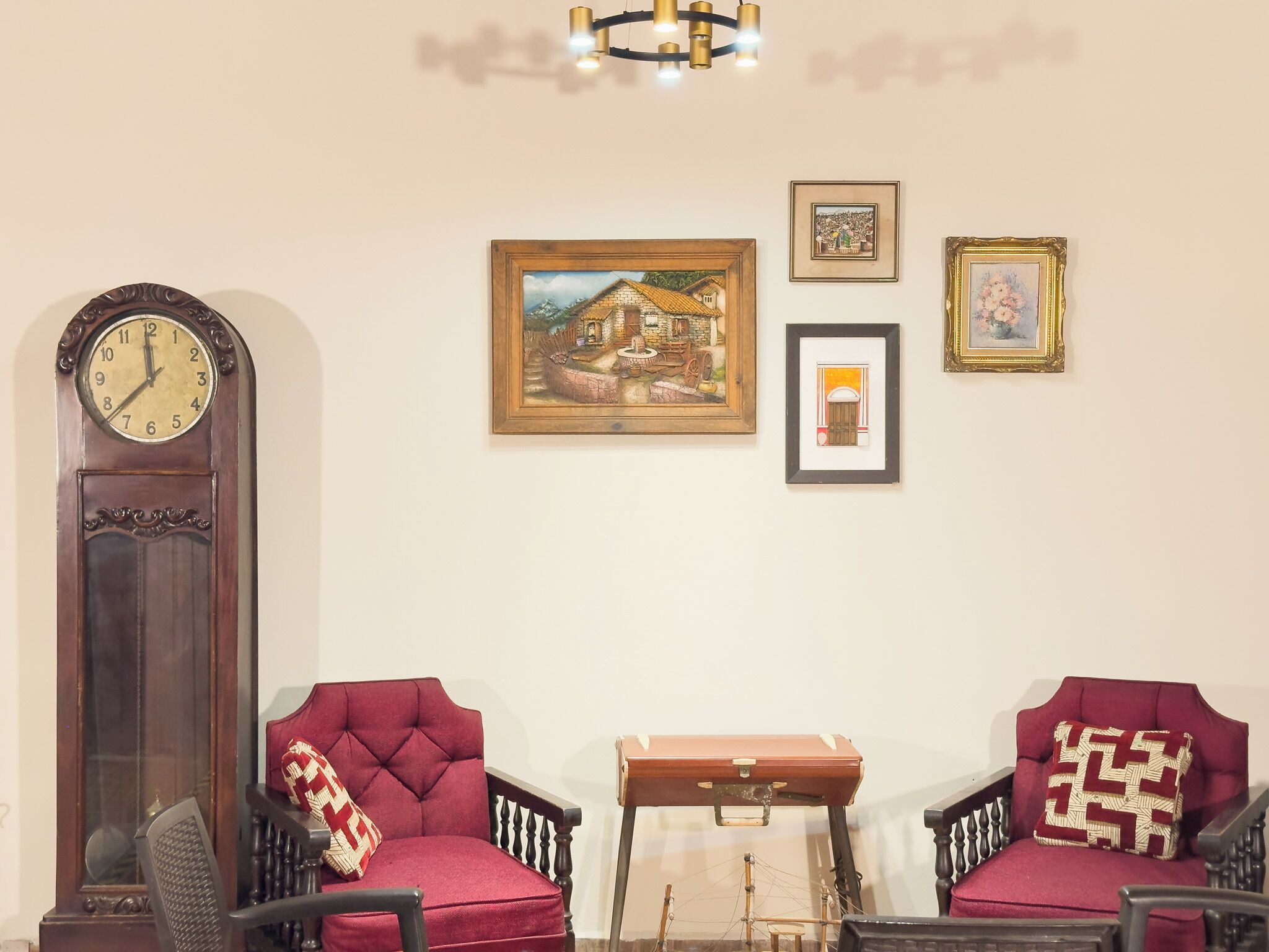A room with a grandfather clock, two red upholstered chairs, a wooden chest, and three framed pictures on the wall.