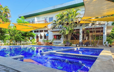 A swimming pool with a tiled edge, surrounded by lounge chairs and umbrellas, with a building in the background.