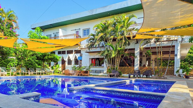 Una piscina con borde de azulejos, rodeada de sillones de descanso y sombrillas, con un edificio al fondo.