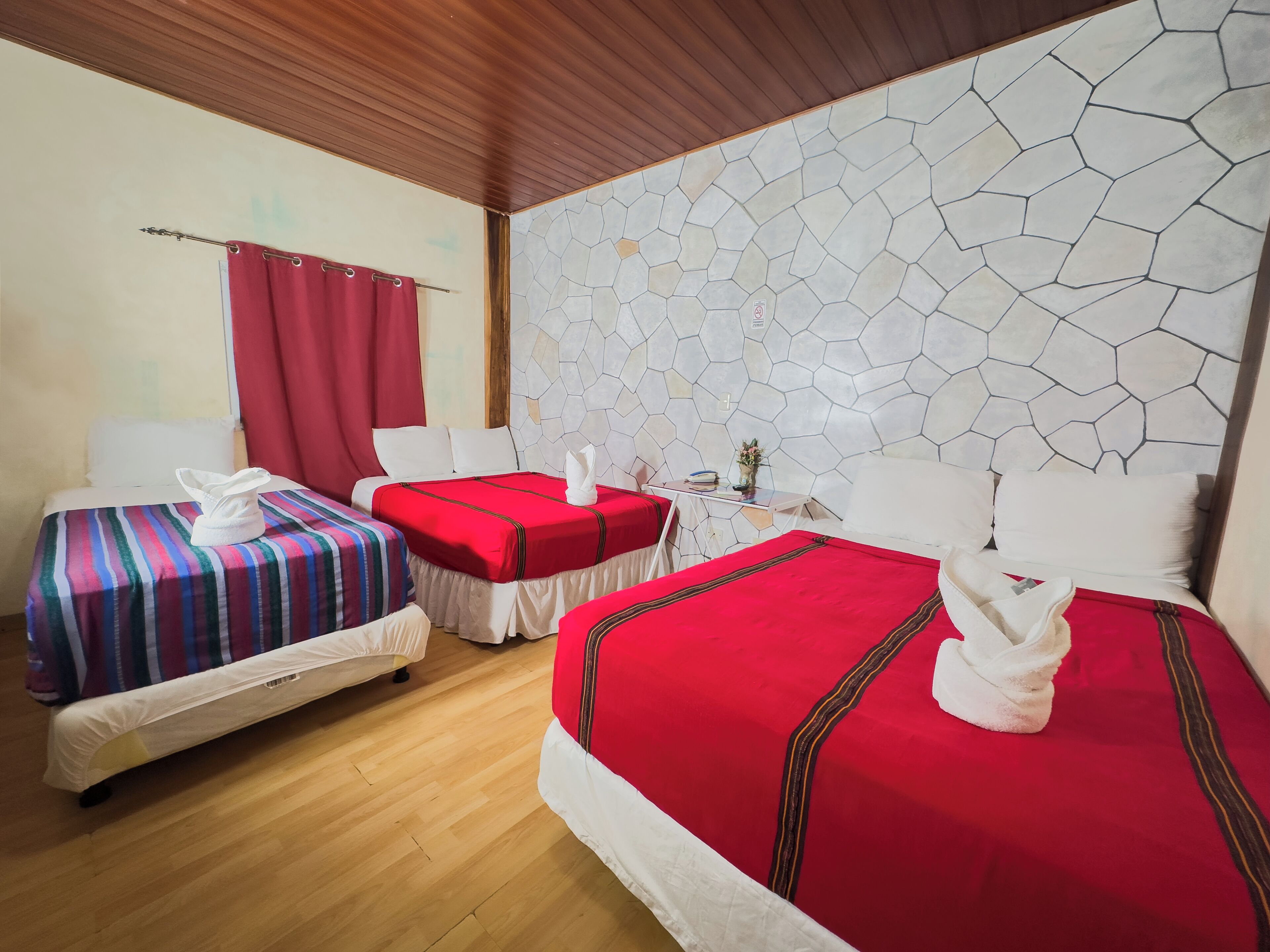 A hotel room with two single beds, a large bed, wooden flooring, a stone-patterned wall, and a small table with a vase.