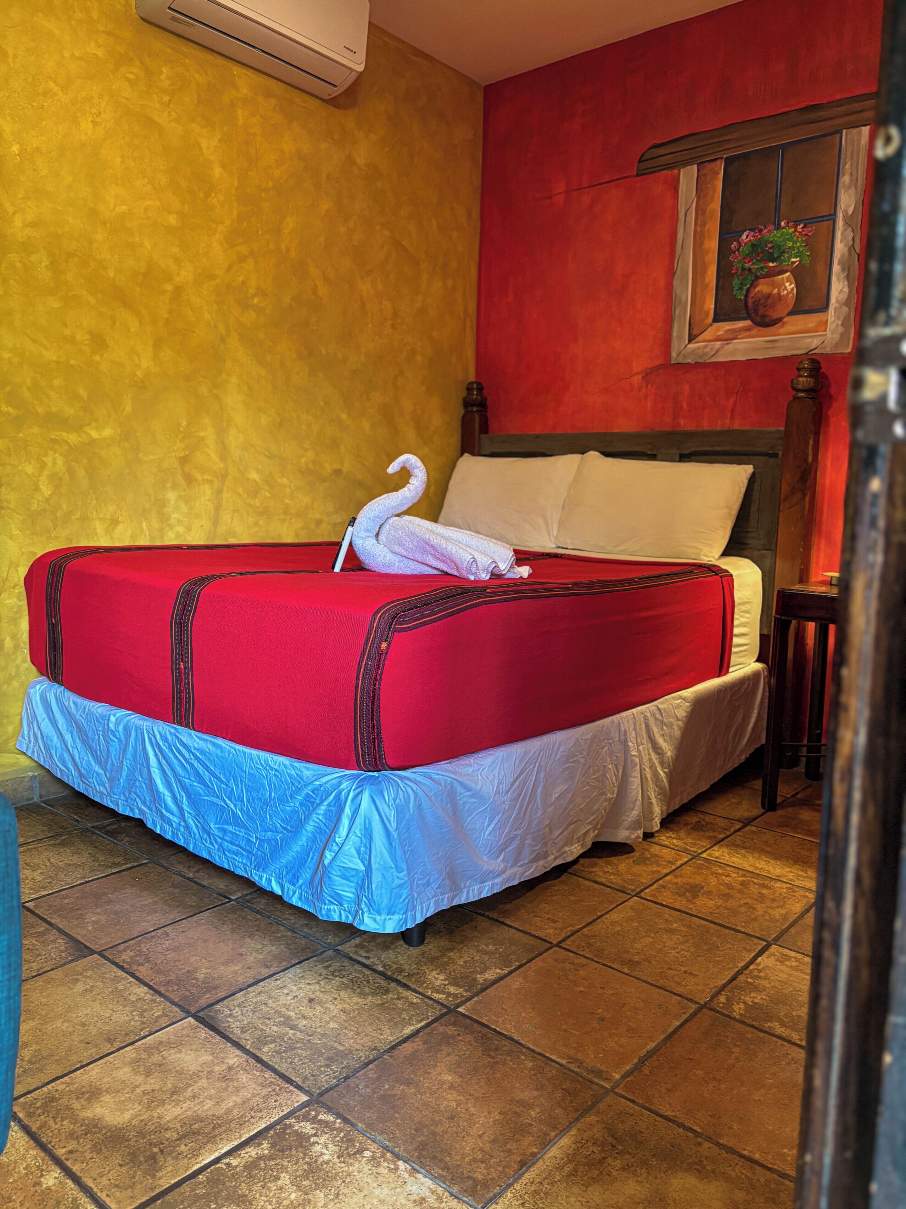 A hotel room with a bed, a red and white bedspread, a blue skirted bed base, a wooden headboard, a red wall, a window with a plant, and a tiled floor.