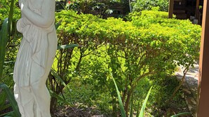 Un jardín con una estatua, abundante follaje y un edificio con techo de madera y luces colgantes.