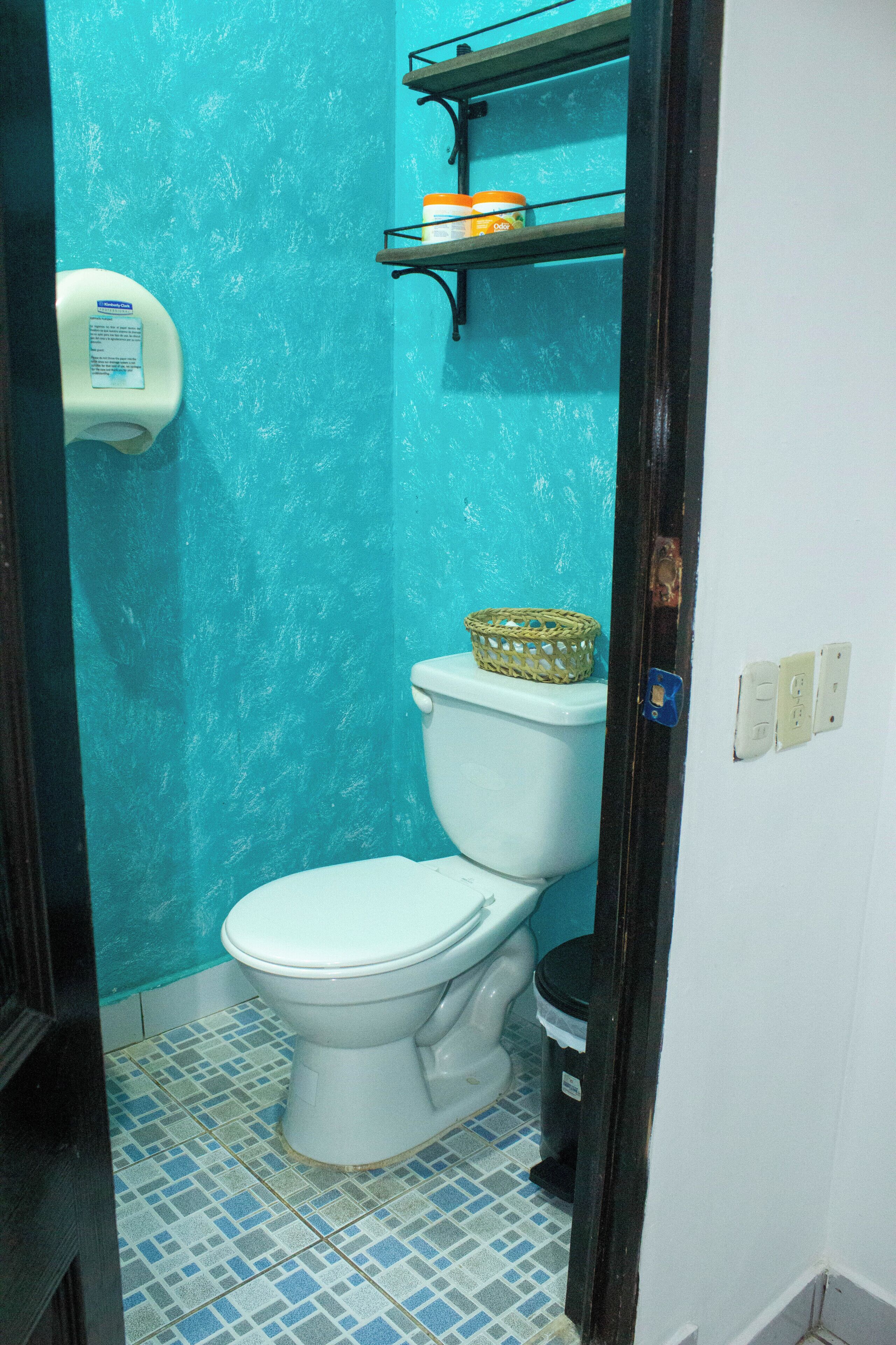 Un baño con inodoro, un cubo de basura, dispensador de jabón y una repisa con una cesta.