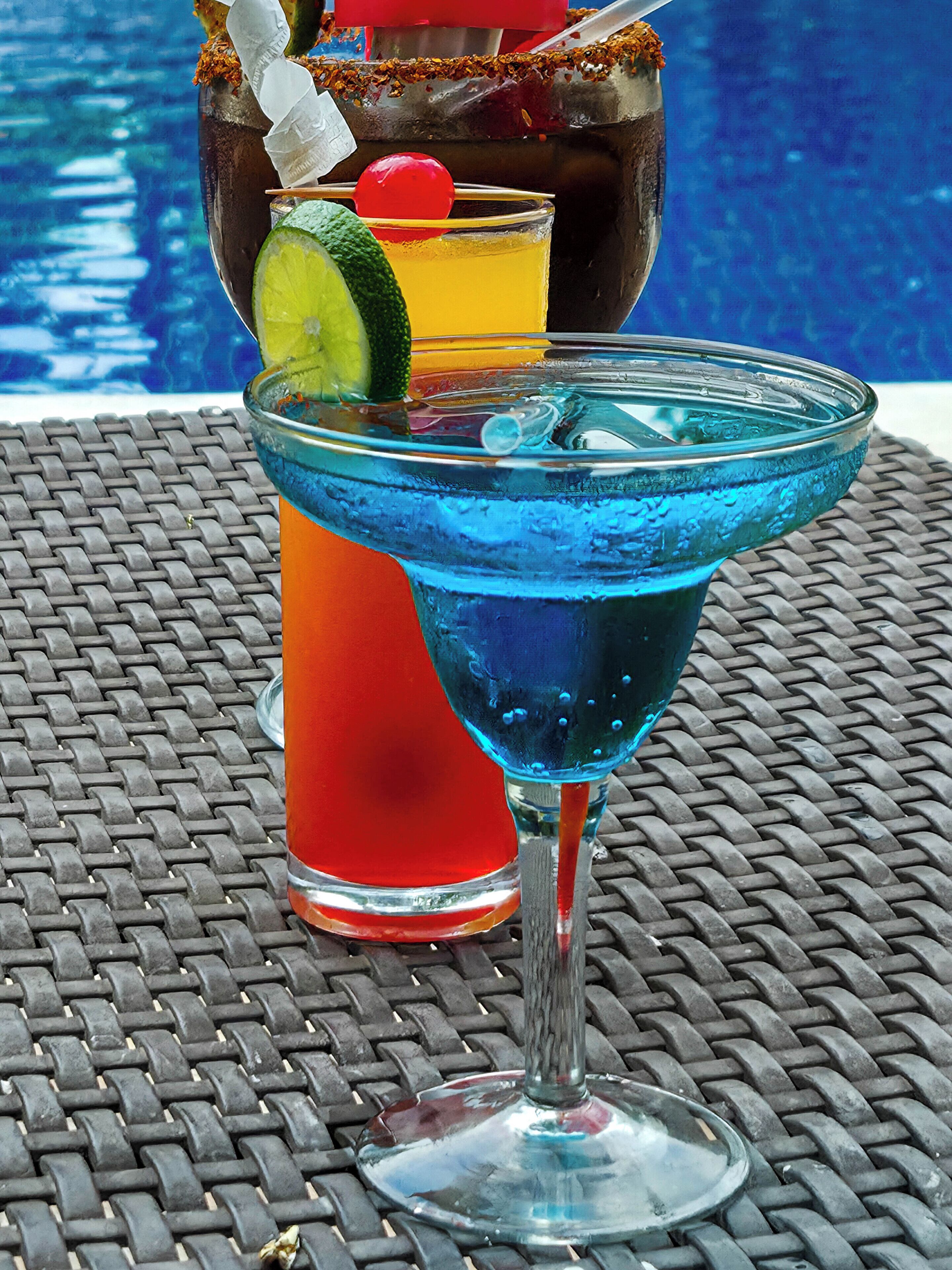 Escena junto a la piscina con una bebida en un vaso azul, un trago rojo en un vaso alto y una botella decorativa con pajita.
