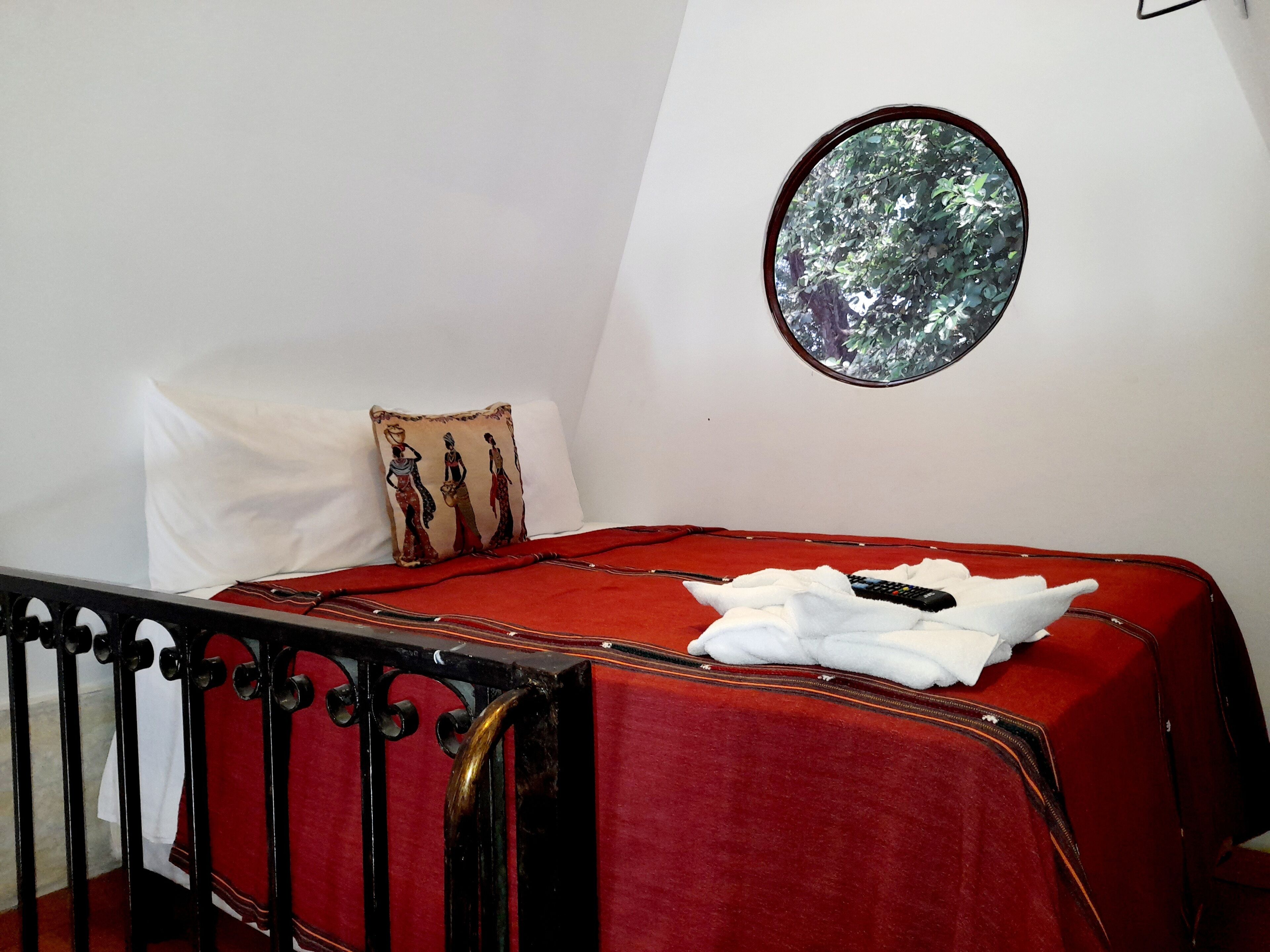 A bed with a red cover, a white pillow, a decorative pillow with a tree design, a remote control, and a circular mirror on the wall.