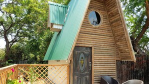 Una casa de madera con techo verde, un ventanal redondo y una puerta con diseño decorativo. Se observa una cerca de madera y plantas en macetas al frente.