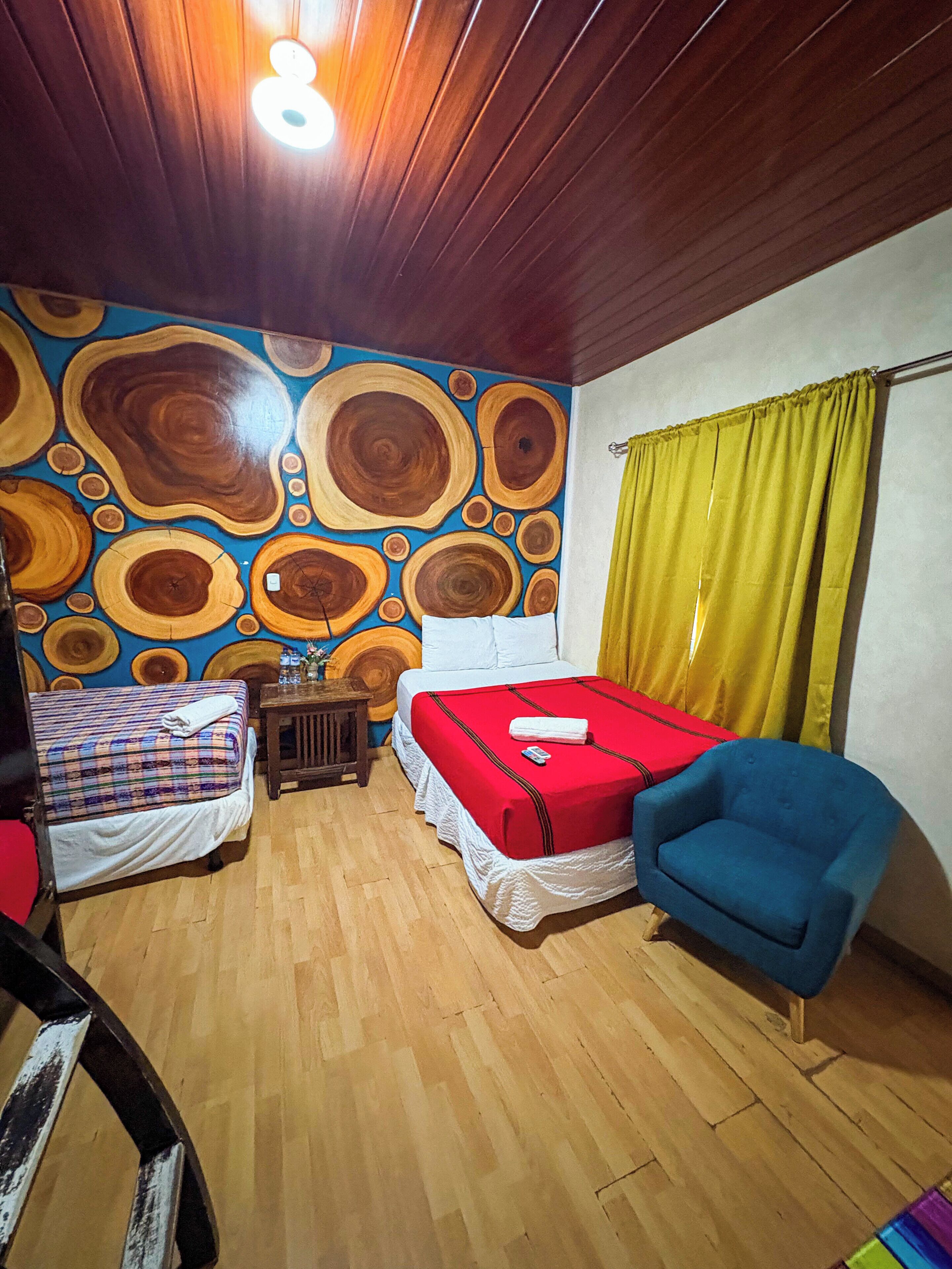 Habitación con dos camas, una silla azul, piso de madera y una pared con diseño circular.