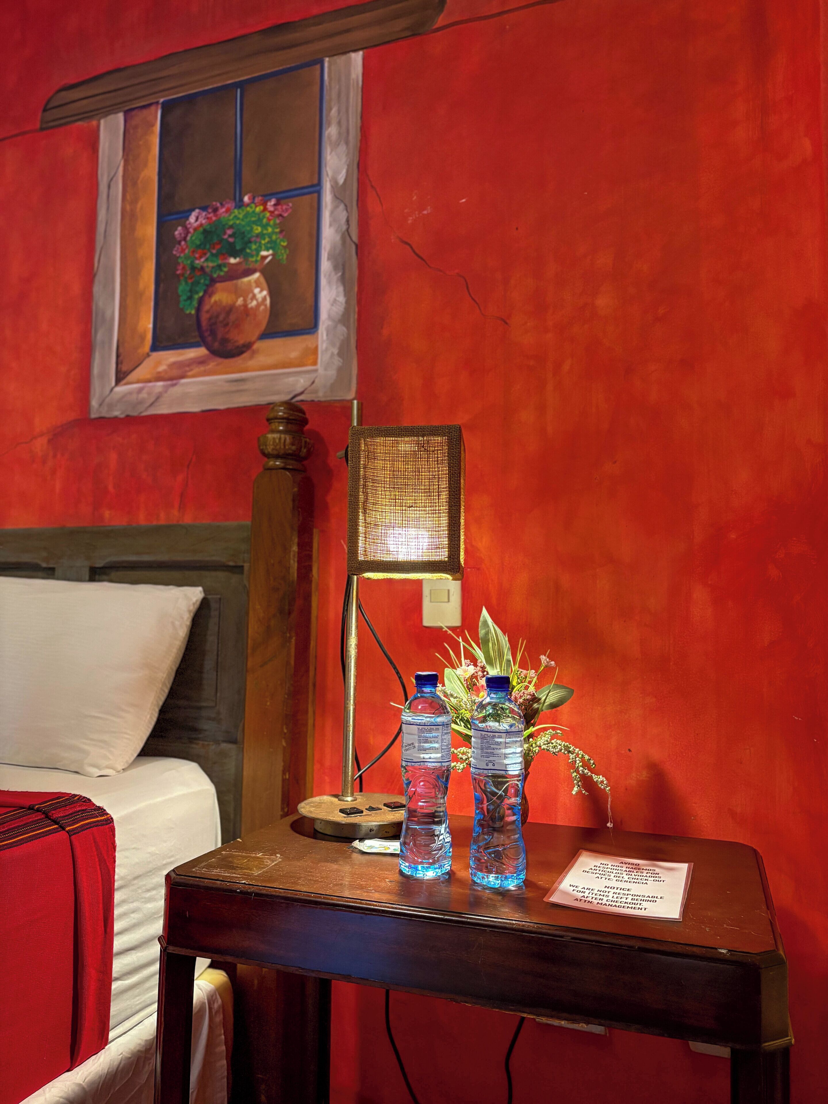 A hotel room with a bed, a small table with water bottles and a plant, a lamp, and a painting on the wall.
