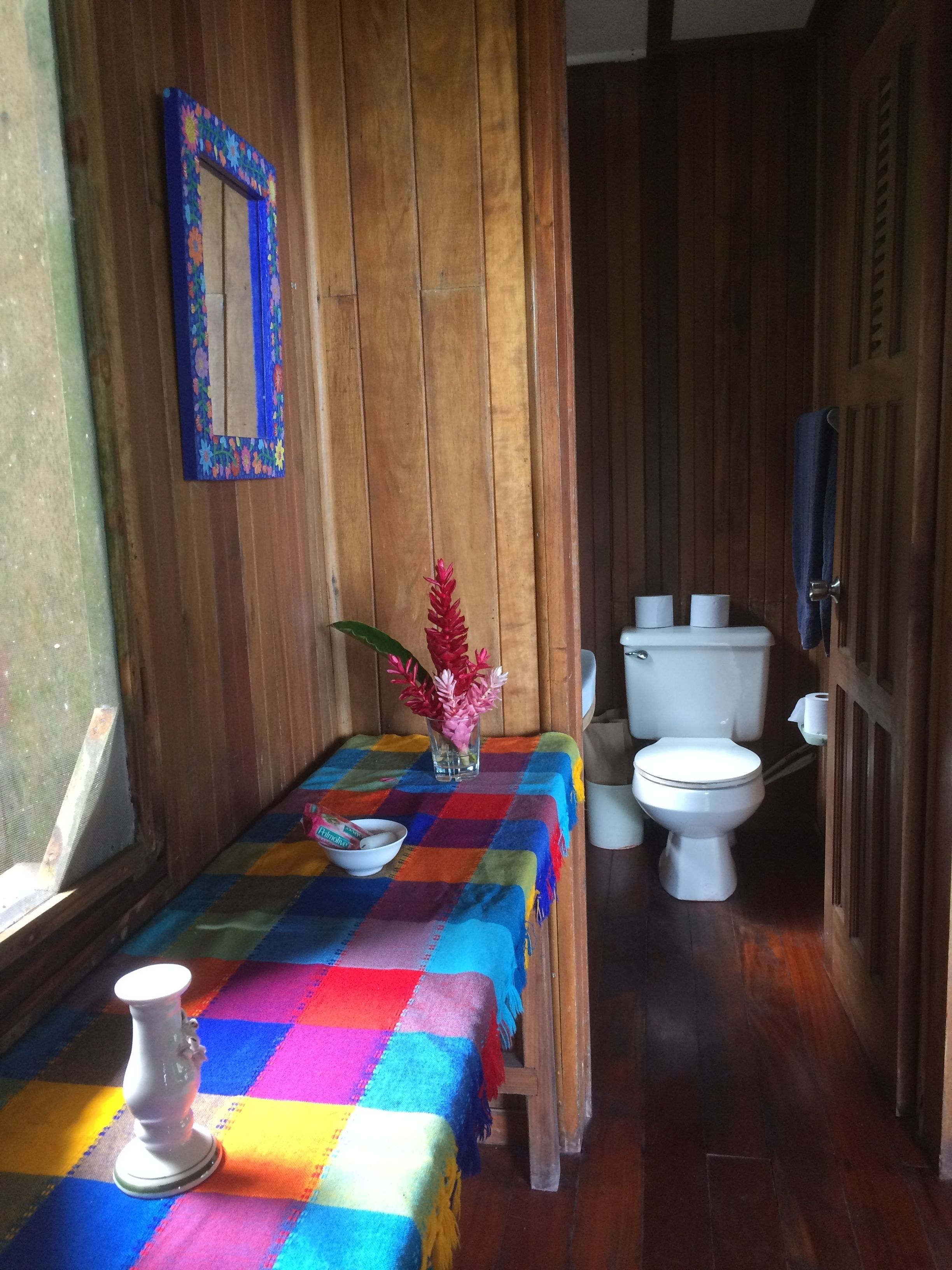 Un baño con piso de madera, un mantel a cuadros de colores, un florero blanco con flores, un inodoro blanco y un espejo con marco azul.