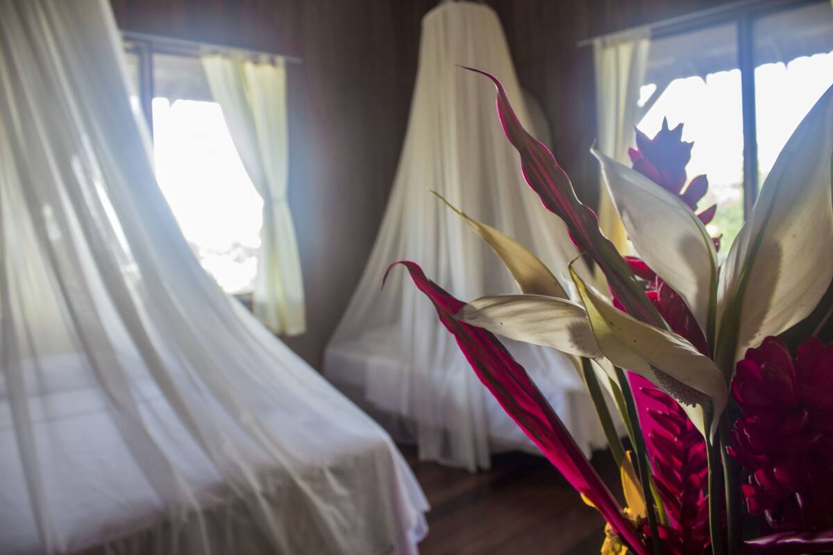 A room with a bed covered in white sheets, a window with sheer curtains, and a vase with red flowers.