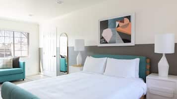 Ein modernes Hotelzimmer mit einem großen Bett, einem blaustürkisen Sessel, einem Nachttisch mit Lampen und einem großen Fenster mit Blick nach draußen.