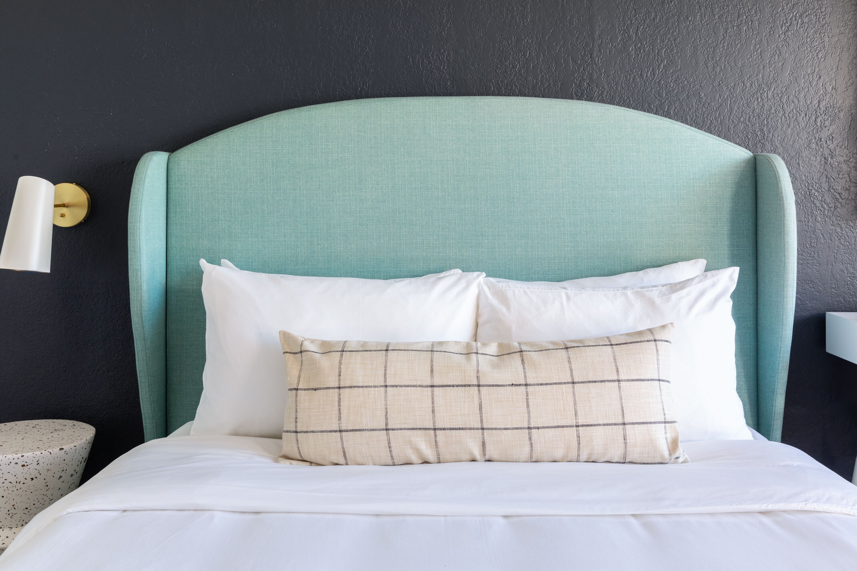 Una cama con cabecera color turquesa, ropa de cama blanca y almohada de cuadros.