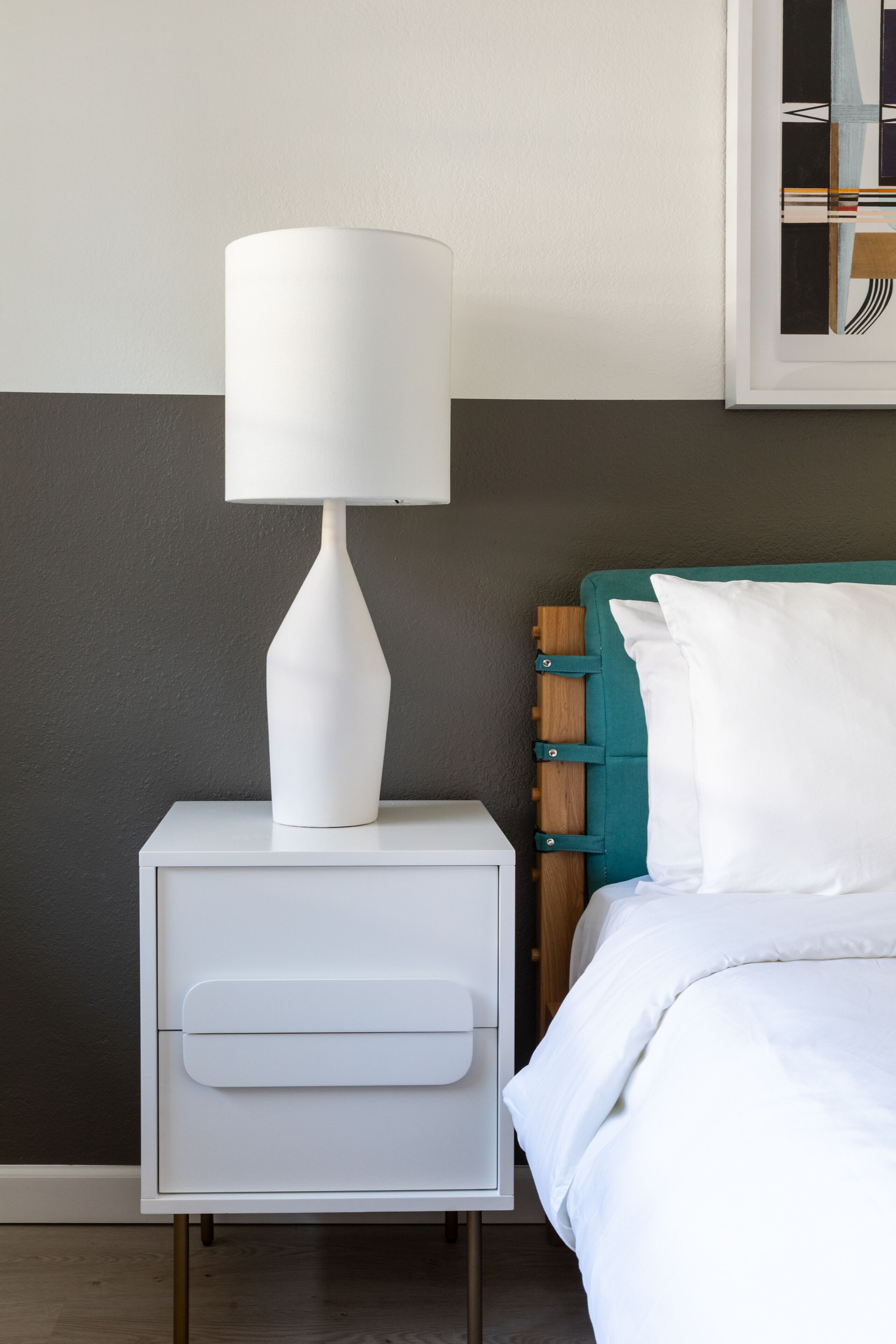 A white bedside table with a lamp, a teal headboard, and a white bed with pillows.