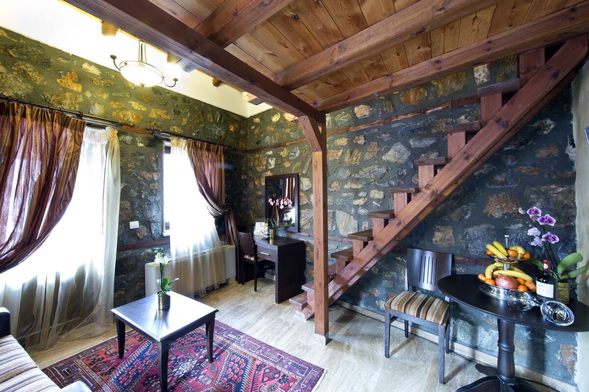 A rustic room with a stone wall, wooden staircase, and a dining table with fruit and flowers.