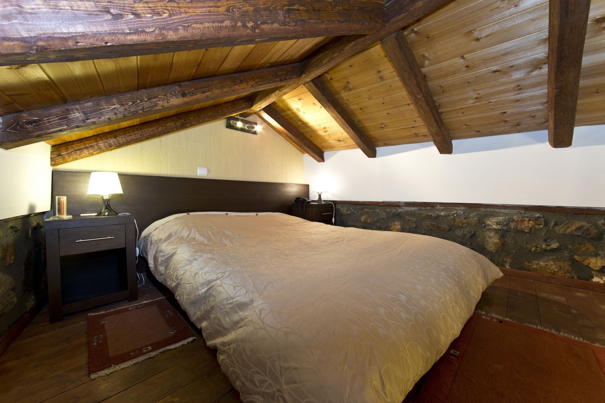 Un dormitorio con techo de madera, cama con manta color beige, mesita de noche con lámpara y pared de piedra.