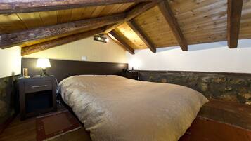 Un dormitorio con techo de madera, cama con manta color beige, mesita de noche con lámpara y pared de piedra.