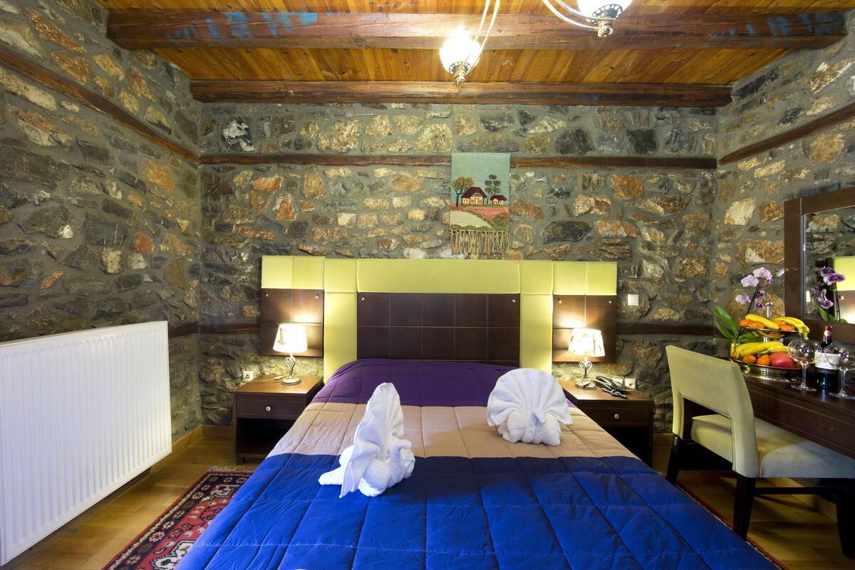 A stone-walled bedroom with a wooden ceiling, a bed with two towels, bedside lamps, a chair, and a fruit bowl.