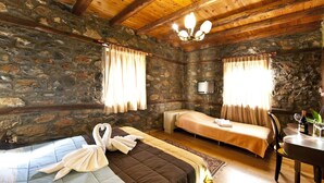 Una habitación rústica con paredes de piedra, vigas de madera, una cama con toalla con forma de cisne, un pequeño sofá, una mesa con vasos y una botella, y una lámpara en el techo.