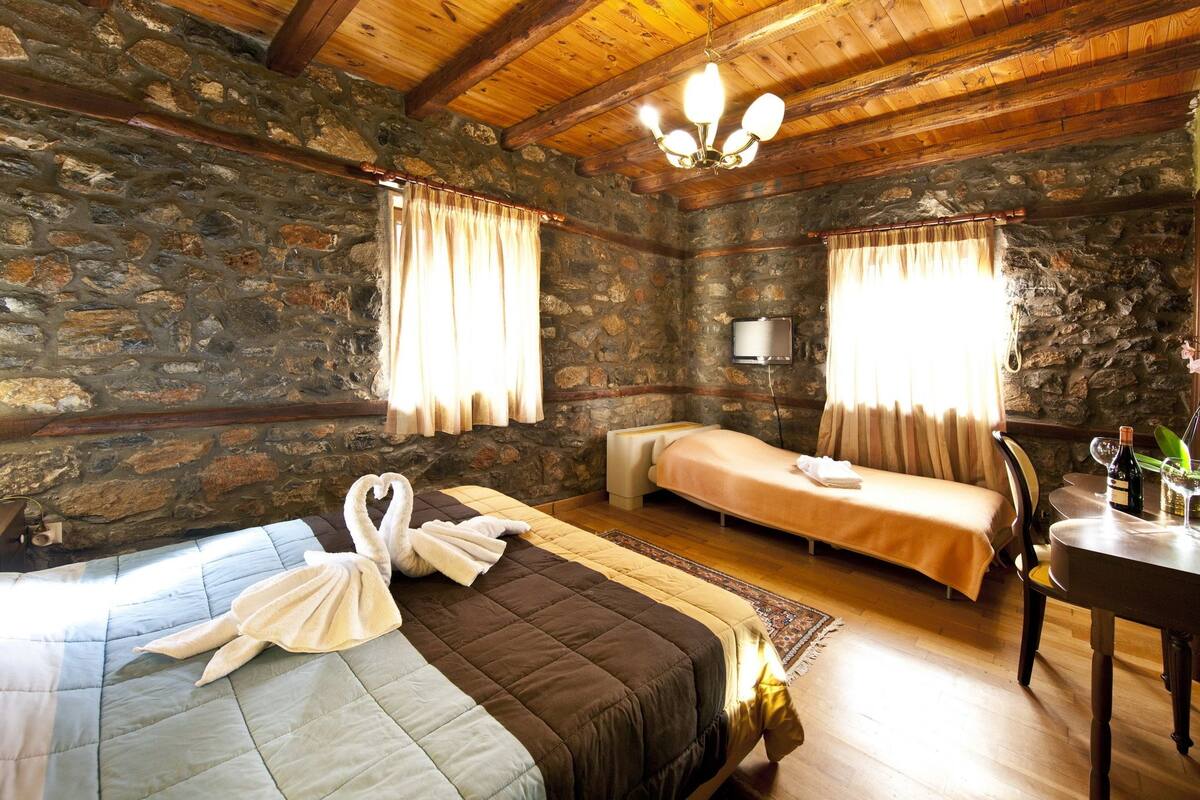 A rustic room with stone walls, wooden beams, a bed with a swan-shaped towel, a small sofa, a table with glasses and a bottle, and a ceiling light.