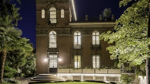 A large, ornate building with multiple windows and balconies, surrounded by lush greenery and a well-lit entrance. - Villa Liberty - Lake Como Rooms & SPA (Como)