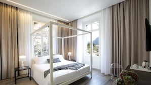 A modern hotel room with a four-poster bed, a small bedside table with a lamp, a desk with a chair, and a large window offering a view of the outdoors. - Villa Liberty - Lake Como Rooms & SPA (Como)