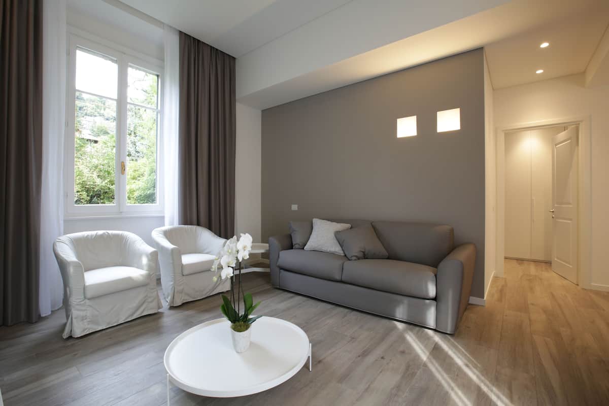 A modern living room with a grey sofa, two white armchairs, a white coffee table with a plant, and a large window with curtains.