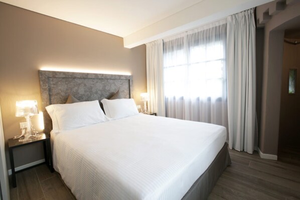 Una habitación de hotel con una cama grande, un cabecero con lámpara, dos mesitas de noche con lámparas y una ventana con cortinas.