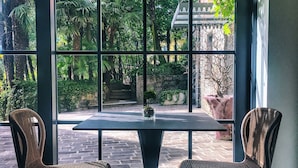 A table with two chairs in front of a large window with a view of greenery and a building. - Villa Liberty - Lake Como Rooms & SPA (Como)