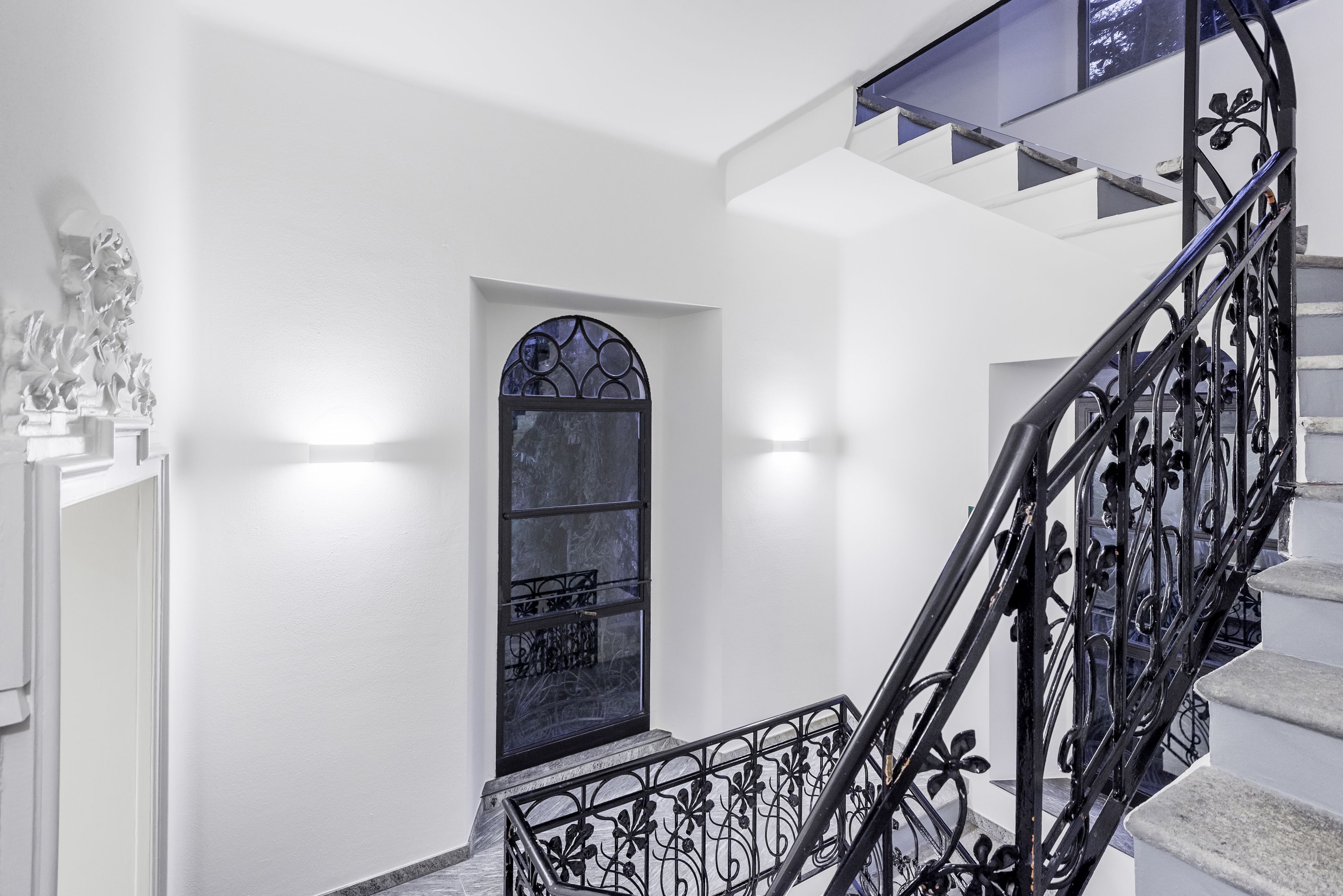 Una escalera con barandilla de hierro forjado negro y pared blanca con un ventanal decorativo.