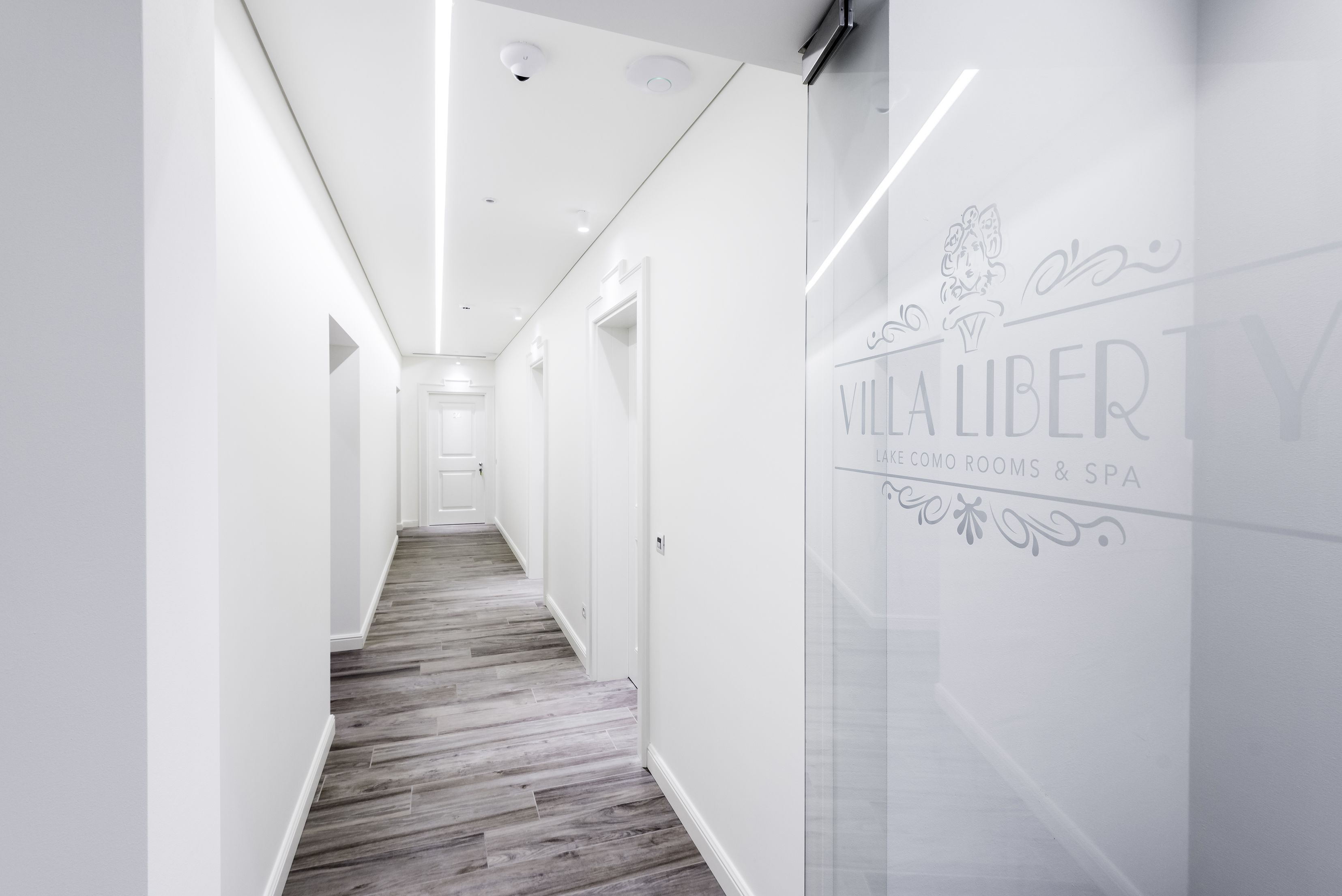 Een moderne hal met een houten vloer, witte muren en een glazen wand met de tekst 'Villa Libre Lake Como Rooms & Spa'.