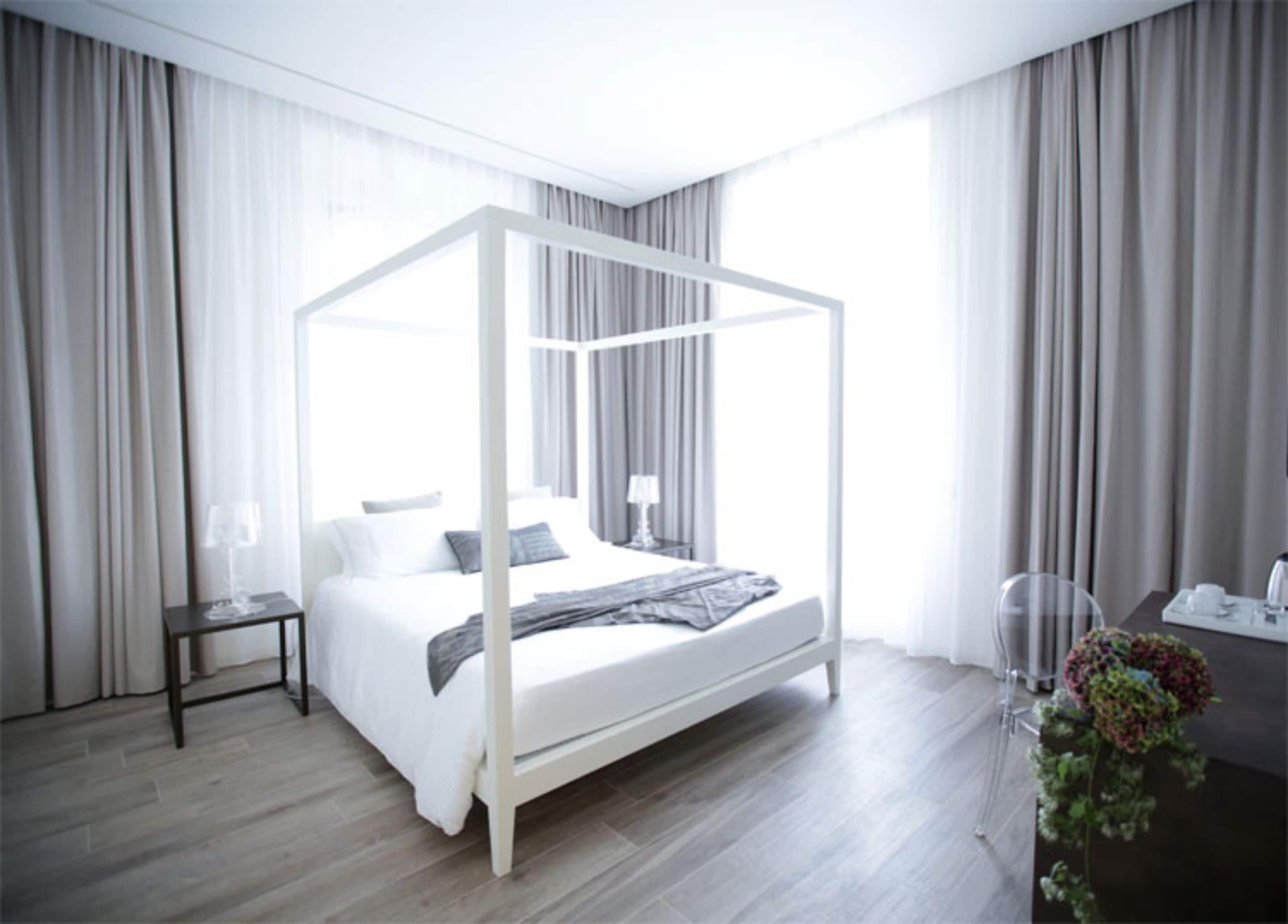 A modern bedroom with a four-poster bed, a bedside table with a lamp, and a desk with a vase of flowers.